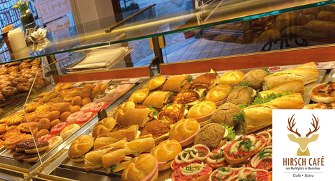 A selection of delicious baked goods and snacks in a glass showcase. In the background, fresh rolls and various sandwiches can be seen.