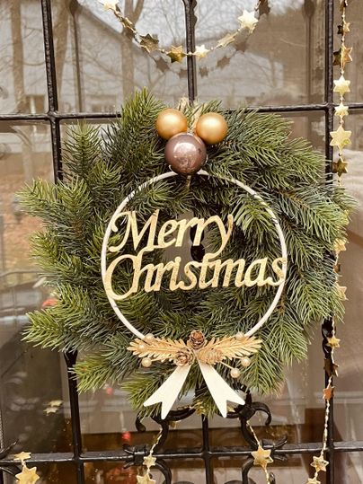 Ein Weihnachtskranz hängt an einer Tür. Er ist mit einer goldenen „Merry Christmas“-Botschaft und dekorativen Kugeln geschmückt.