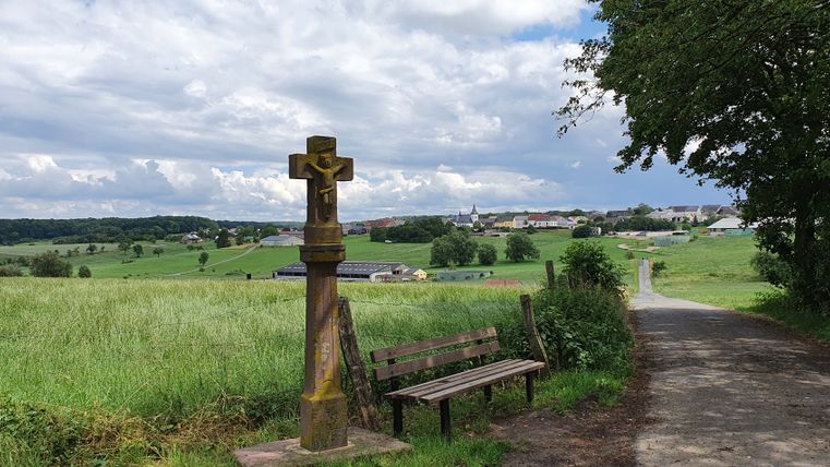Croix de chemin et banc avec vue sur Mötsch, entourés de champs verts et d'un ciel nuageux.