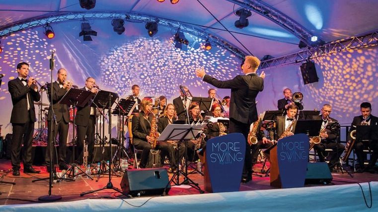 Eine große Band spielt auf einer Bühne unter einem Zelt. Der Dirigent leitet die Musiker, während die Lichter im Hintergrund leuchten.