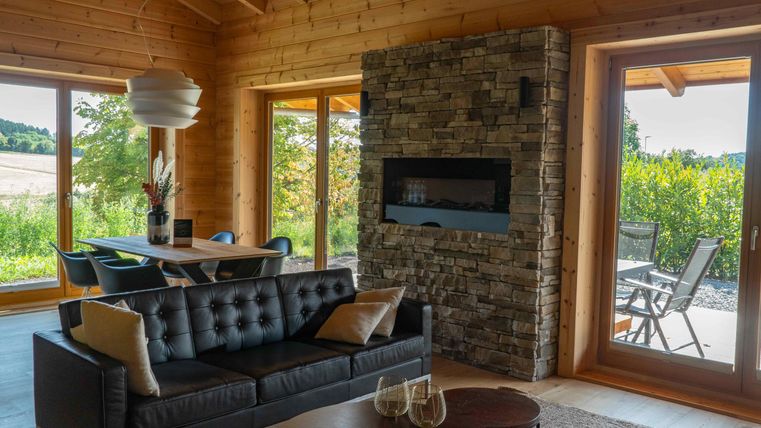 Eine moderne Wohnraum mit einem schwarzen Sofa und einem gemütlichen Steinkamin. Große Fenster bieten einen Blick auf die grüne Umgebung.