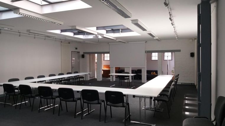 Een heldere vergaderzaal met lange tafels en stoelen. Grote vensters zorgen voor veel natuurlijk licht.