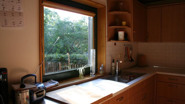 Een lichte keuken met houten kasten en een groot raam. Buiten is er een groene tuin te zien met een hek.