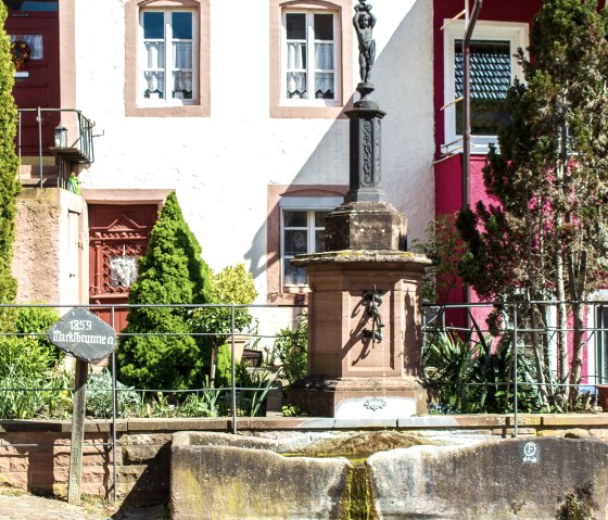 Une fontaine historique datant de 1859 se trouve devant un bâtiment aux façades rouges et blanches à Dudeldorf. Elle est entourée de plantes et d'une balustrade., © Tourist-Information Bitburger Land_M. Mayer
