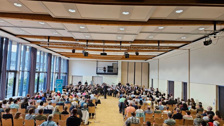 Ein großer Raum mit vielen Sitzplätzen ist mit Publikum gefüllt. Auf der Bühne spielt ein Orchester vor einer großen Zuschauermenge.