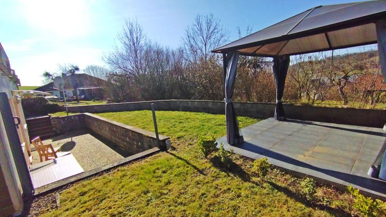 Un beau jardin avec une terrasse et un pavillon couvert. À l'arrière-plan, on peut voir des arbres et un ciel dégagé.
