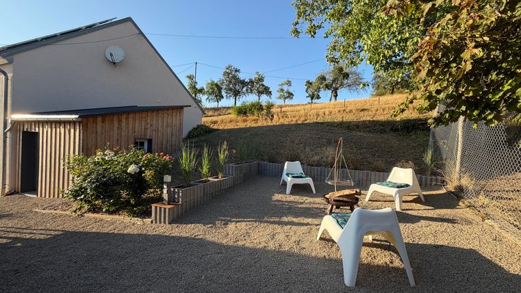 Een gezellige buitenruimte met een terras en witte stoelen. Op de achtergrond zijn bomen en een zachte helling zichtbaar.