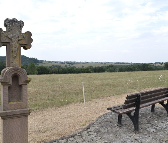 Bench and stone cross in a rural landscape with sweeping views over fields and hills., © TI Bitburger Land