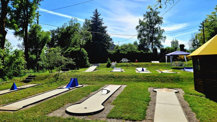 Eine MiniGolf-Anlage mit 18 Bahnen und grünen Wiesen. Im Hintergrund stehen Bäume und ein sonniger Himmel. Sitzmöglichkeiten unter schattenspendendem Pavillons oder Baumkronen.