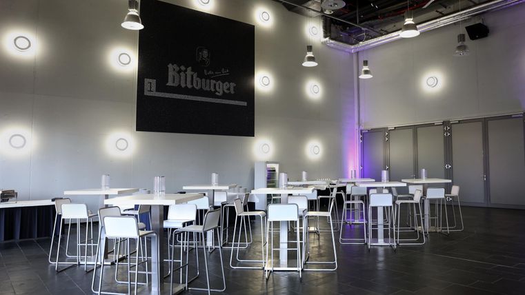 A modern restaurant with high tables and chairs. The walls are kept simple, with soft lights and a large Bitburger logo.