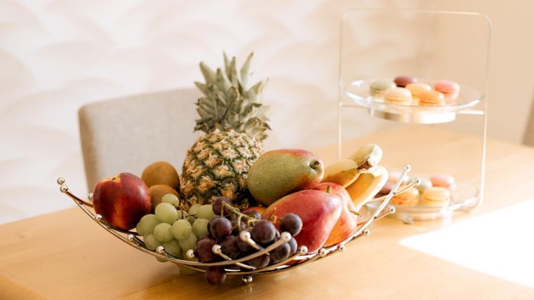 A stylish fruit bowl with apples, grapes, a pineapple, and a pear is on a table. Behind the bowl, there is a stand with macarons.