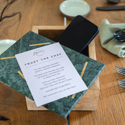 A stylish table with a wooden tabletop, on which a green menu and a smartphone lie. Next to the menu, cutlery and napkins are decoratedly arranged.
