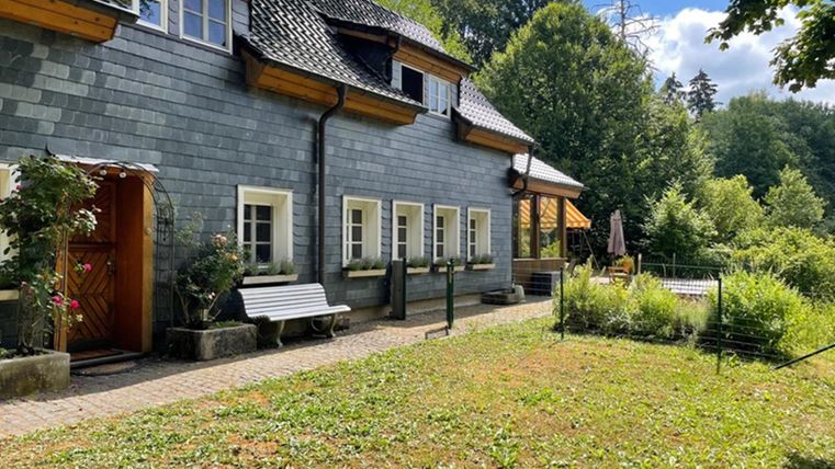 A charming, gray house with a beautiful garden. In the background, trees and a sunny landscape can be seen.