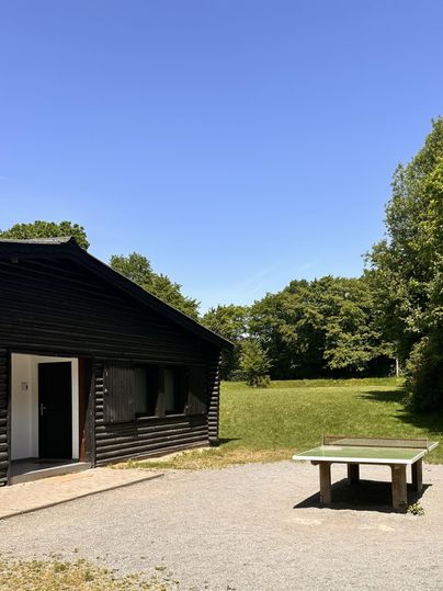 Een zwart geschilderd houten huis staat voor een heldere blauwe lucht. Naast het huis staat een tafel voor tafeltennis op een groene wei.