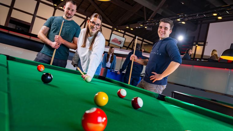 Trois jeunes personnes se tiennent à la table de billard et sourient. Des boules de billard colorées sont posées sur la table.
