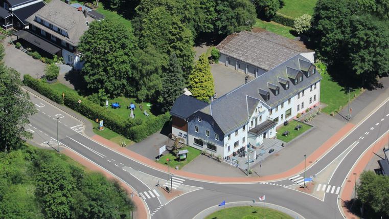 Een vogelperspectief op een rotonde met een wit gebouw en veel groen eromheen. Je ziet ook enkele auto’s en paden in de afbeelding.