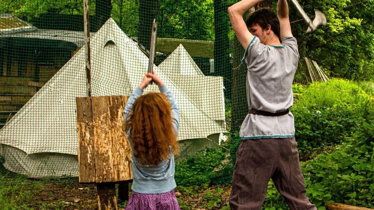 Ein Kind und ein Erwachsener üben das Werfen von Äxten. Im Hintergrund sind ein Zelt und Bäume zu sehen.