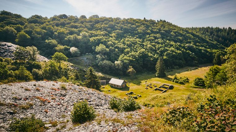 Vue sur la prairie des hommes et la maison à refendre nouvellement construite. Sur la gauche, on voit beaucoup d'ardoise et, en arrière-plan, un paysage de forêt.