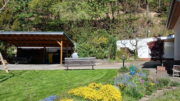 Ein schöner Garten mit bunten Blumen und einer grünen Wiese. Im Hintergrund sind Bäume und eine überdachte Terrasse sichtbar.