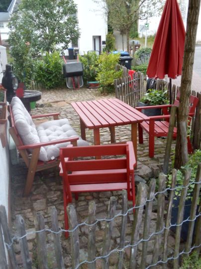 Eine gemütliche Sitzecke im Freien mit roten Möbeln und einem Sonnenschirm. Umgeben von Pflanzen und einem kleinen Zaun.