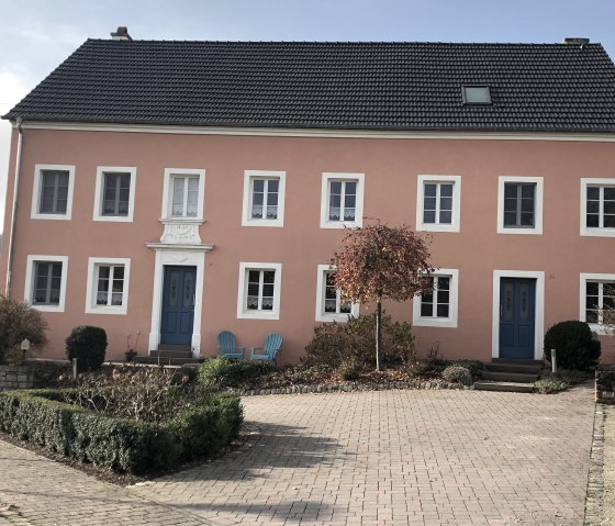 An old farmhouse with a pink façade and blue doors in Oberweis. In front of it are two blue chairs and a small tree in the front garden., © TI Bitburger Land - Steffi Wagner