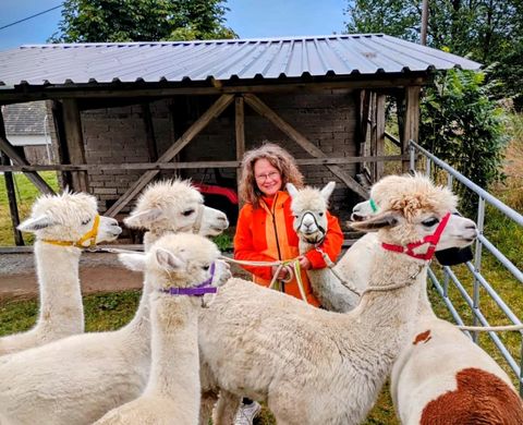 Eine Frau steht in der Mitte einer Gruppe von Lamas. Im Hintergrund ist ein Stall zu sehen.