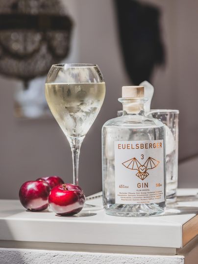 A glass with a clear drink is standing next to a bottle of EUELSBERGER GIN. Two red apples are lying on the table.