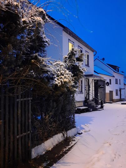 Eine verschneite Straße bei Dämmerung mit einem weißen Haus und schneebedeckten Bäumen. Die perfekte Winterlandschaft vermittelt eine ruhige Atmosphäre.