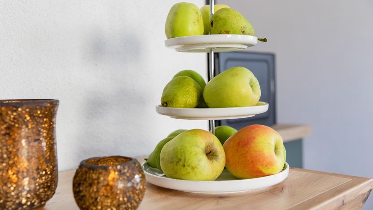 Ein modernes Obstregal mit grünen Äpfeln und einer roten Birne. Im Hintergrund sind dekorative Gläser sichtbar.