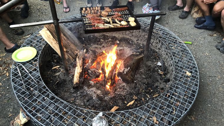 Un feu de camp avec un grill sur lequel différents plats sont préparés. Au fond, des gens sont rassemblés et profitaient de l'ambiance du camping.