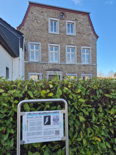 A historic building with many windows and a red roof. In the foreground is an information sign that tells more about the history of the house.
