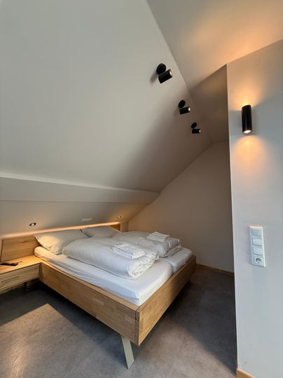A modern bedroom with a wooden bed and fresh bedding. The walls are light, and the lighting is warm and inviting.