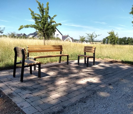 Drei Holzbänke stehen auf einem gepflasterten Platz in einer ländlichen Umgebung. Im Hintergrund sind Bäume, Wiese und Häuser zu sehen., © TI Bitburger Land