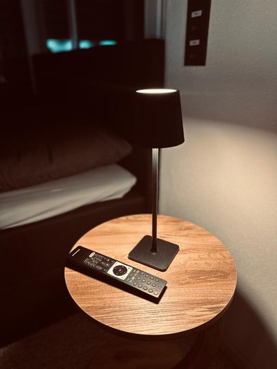 Une table de nuit avec une lampe sur pied et une télécommande. À l'arrière-plan, un lit est visible.