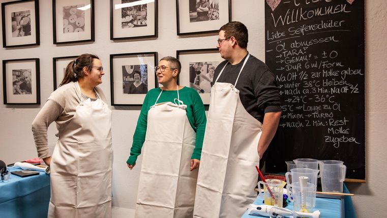 Three people are standing in a room, wearing aprons and laughing together. In the background, pictures are hanging on the wall and there is a board with a welcome message.