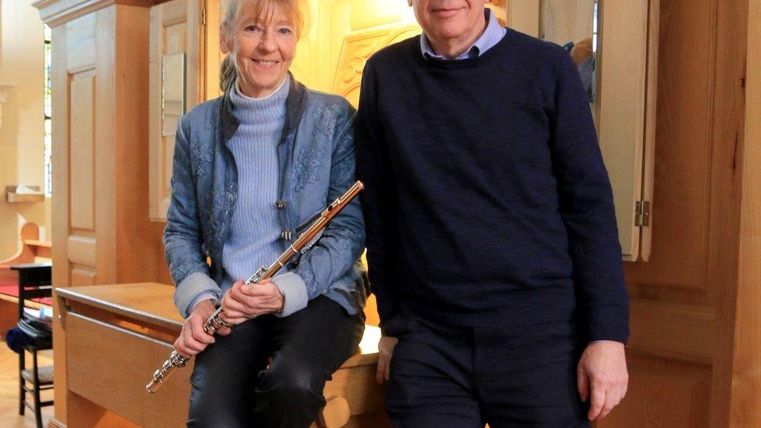 A woman is sitting with a flute in her hand, while a man stands beside her. Both are smiling and posing in front of a musical instrumental background.