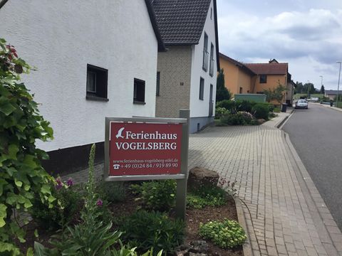 Une plaque de réponse pour une maison de vacances avec l'inscription "Ferienhaus Vogelsberg" se trouve dans une rue calme. Les environs montrent des maisons résidentielles et quelques espèces de plantes.