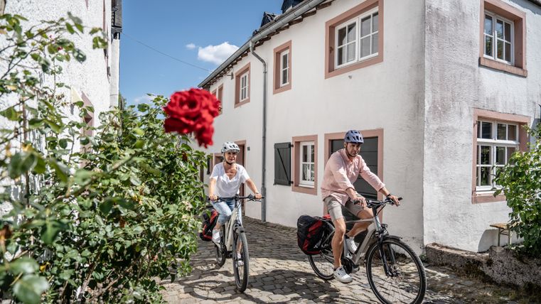 Two cyclists wearing helmets ride through the old town of the historic castle town of Kronenburg in sunny weather. In the left foreground is a rose bush, from which a red rose protrudes into the picture.