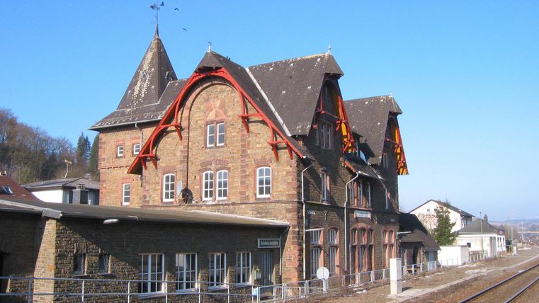 Blick auf die Rückseite des  gemauerten  Bahnhofsgebäudes von den Bahnsteigen. Man erkennt das flache Nebengebäude und das dreistöckige Hauptgebäude mit dem Schriftzug Jünkerath.Im Vordergrund sieht man die Bahngleise.