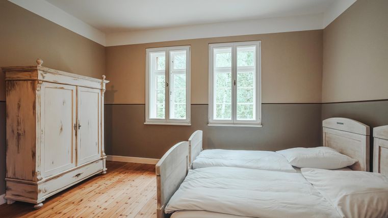 Dit beeld toont een slaapkamer met twee eenpersoonsbedden en een grote antieke kast. Door twee ramen heeft men uitzicht op de bomen buiten.