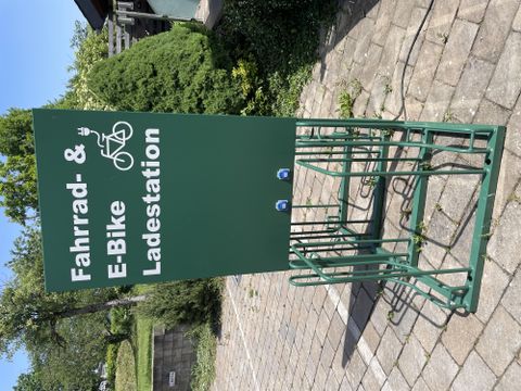 Een fiets- en e-bike oplaadstation staat op een betegeld gebied. Op de achtergrond zijn bomen en struiken zichtbaar.