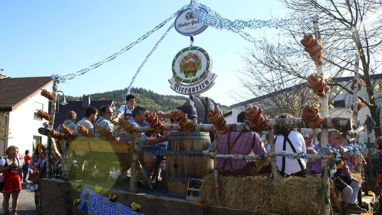 Een feestelijke optochtwagen met vrolijke mensen in traditionele kleding. Er zijn biervaten, pretzels en versieringen te zien die een feestelijke sfeer creëren.