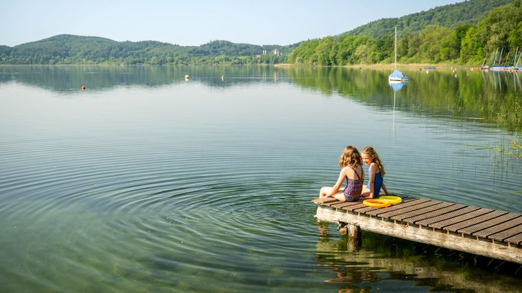 Zwei Kinder sitzen entspannt am Ende eines Holzstegs, der in den ruhigen, klaren Laacher See führt; sie beobachten die kreisförmigen Wellen auf dem Wasser, umgeben von grünen Hügeln und bewaldetem Ufer bei sonnigem Wetter.