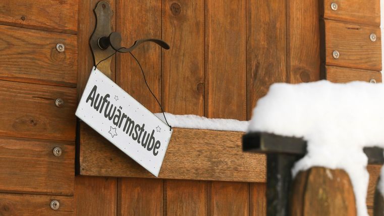 Ein Holzschild mit der Aufschrift "Aufwärmstube" hängt an einer Tür. Schnee liegt auf dem oberen Rand der Tür und auf dem Geländer.