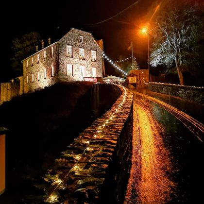 A historic house with illuminated windows stands by a path that gently rises. The path is adorned with lights and the surroundings are nocturnal and calm.
