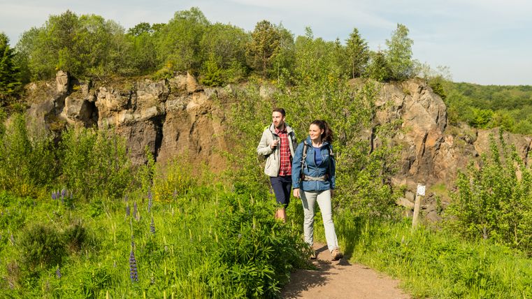 Zwei Wanderer inmitten der Natur auf einem Wanderweg. Im Hintergrund ist eine große Felswand zu sehen. Hierbei handelt es sich um den Vulkan Steffelnkopf.