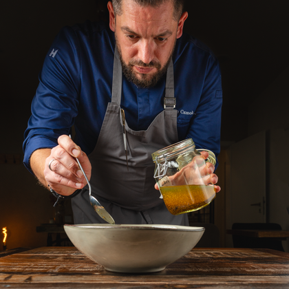 A cook is preparing a meal and adds a sauce from a jar into a bowl. He focuses on the preparation and stands at a wooden table.