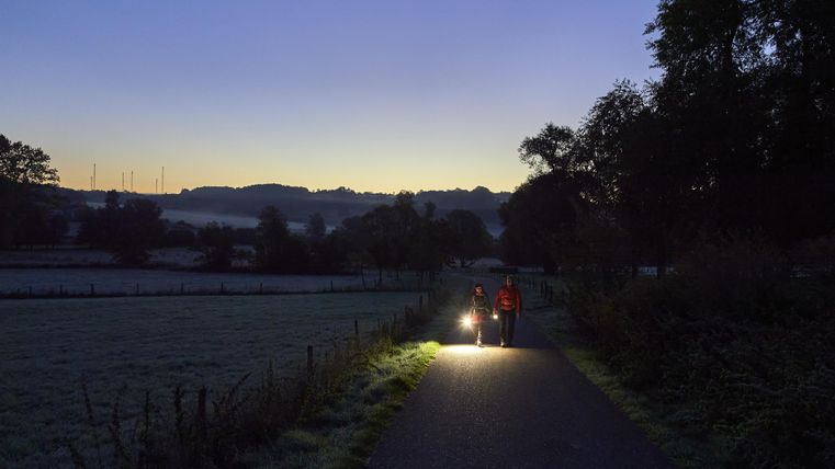 Zwei Personen gehen auf einem Weg in der Dämmerung, während sie eine Taschenlampe tragen. Die Umgebung ist frostig und die Landschaft wirkt ruhig und friedlich.