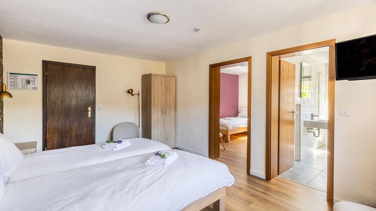 Een lichte kamer met twee eenpersoonsbedden en moderne meubels. Op de achtergrond is een open deur naar een badkamer en een andere slaapkamer zichtbaar.