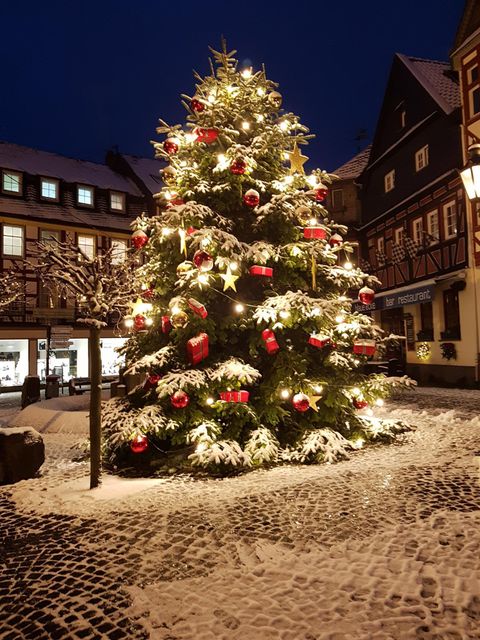 Ein festlich geschmückter Weihnachtsbaum steht in einer verschneiten Stadt. Die Lichter und roten Geschenke verleihen der Szene eine gemütliche Atmosphäre.